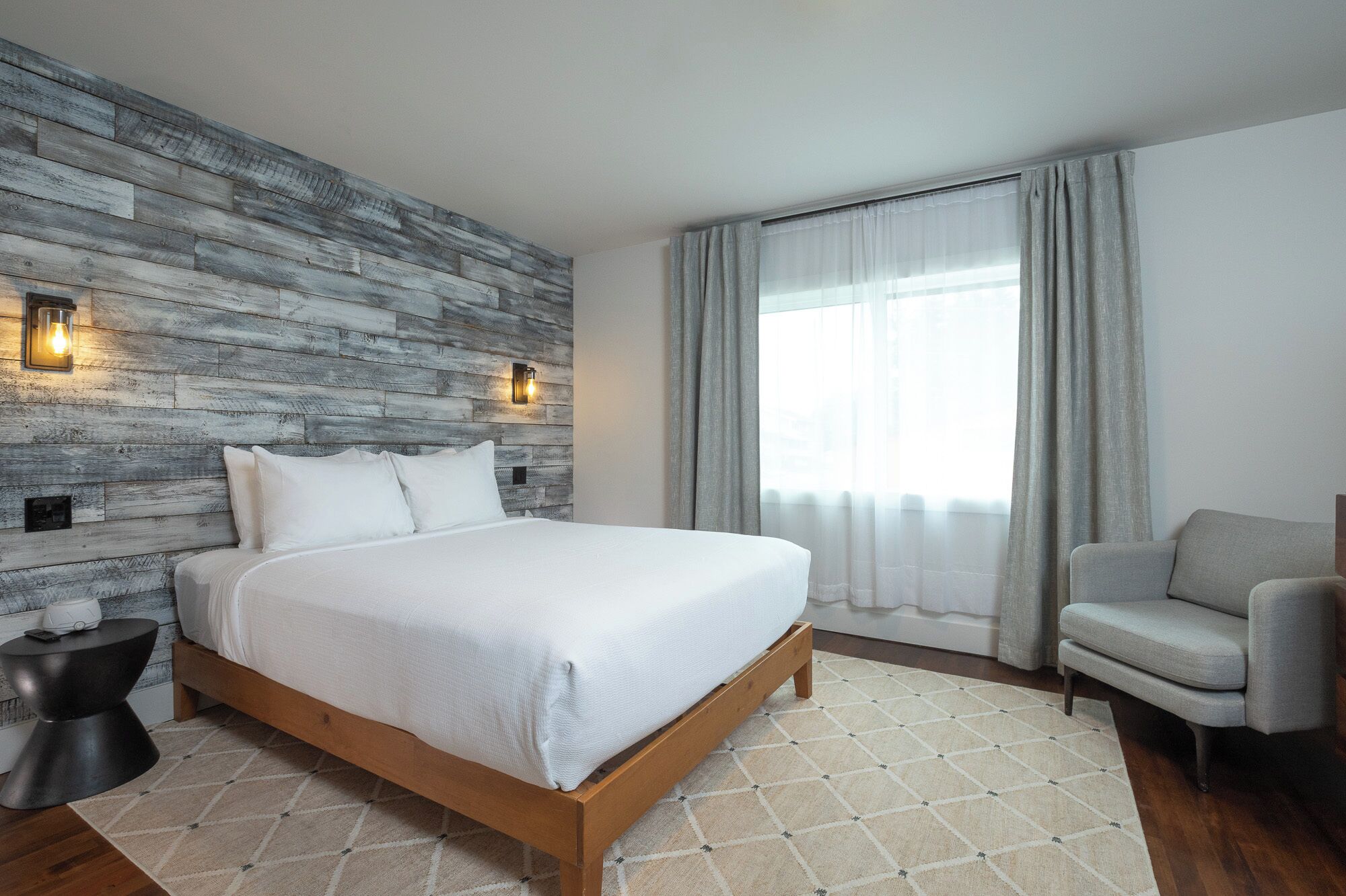 Un dormitorio moderno con cabecera de madera, cama con ropa de cama blanca, mesita de noche, una silla y un ventanal con cortinas.