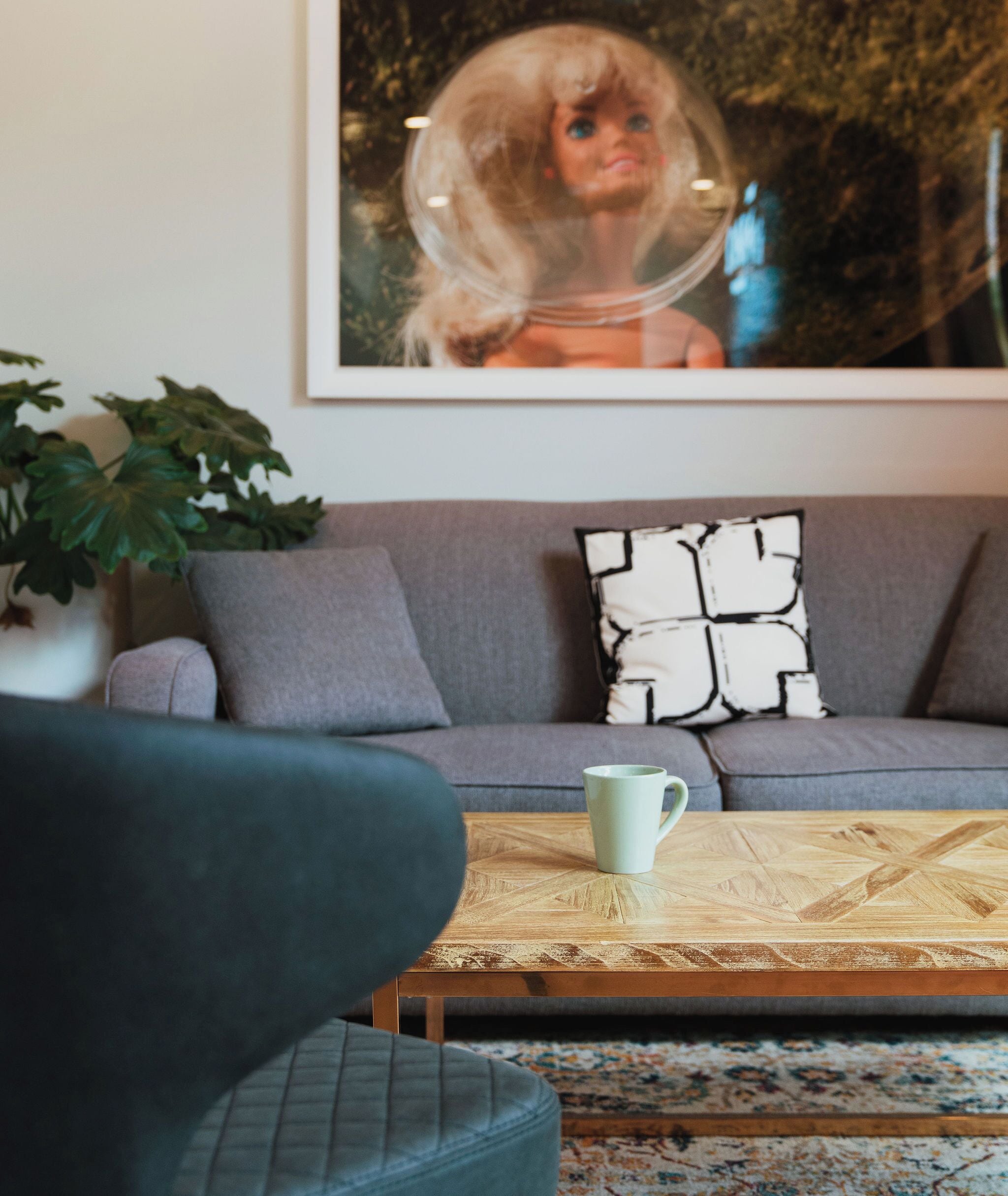 A living room with a grey sofa, a patterned throw pillow, a coffee table with a mug, and a large framed picture on the wall.
