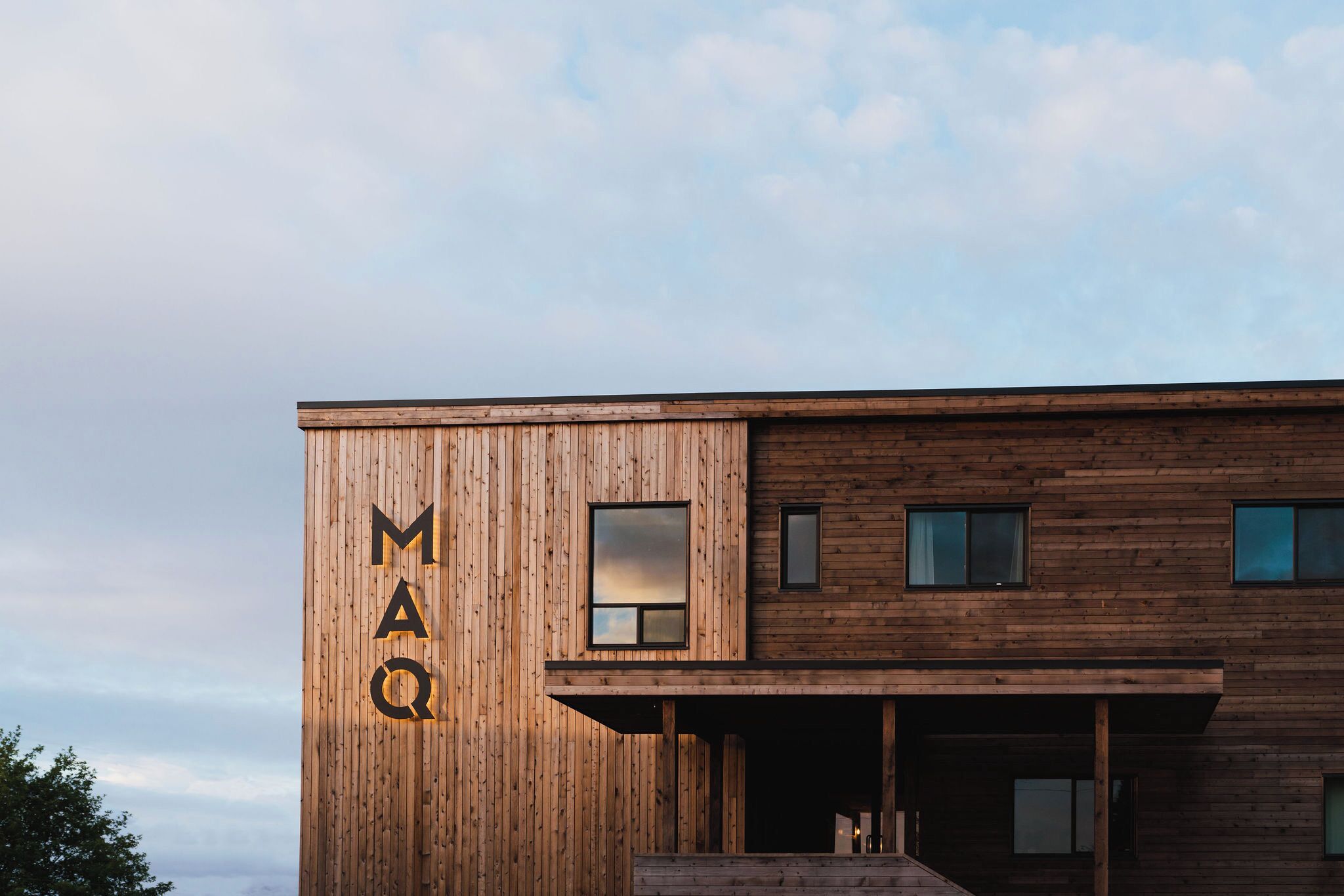 Un edificio moderno con fachada de madera y las letras 'MAQ' iluminadas en el lateral.