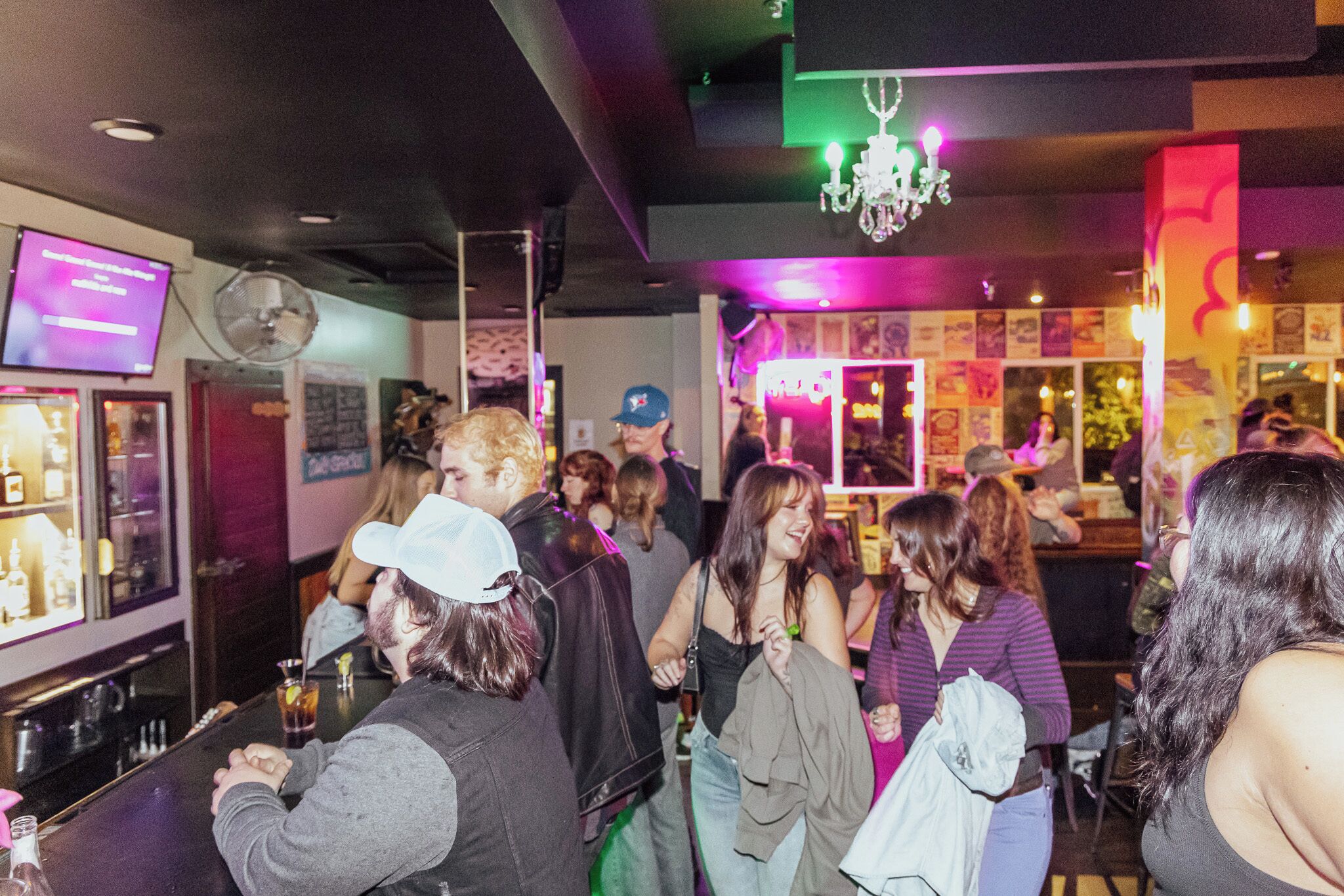 Un bar concurrido donde la gente está de pie y conversando, una pantalla de televisión y una araña.