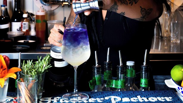 Un cantinero preparando una bebida con varias botellas y una barra al fondo.