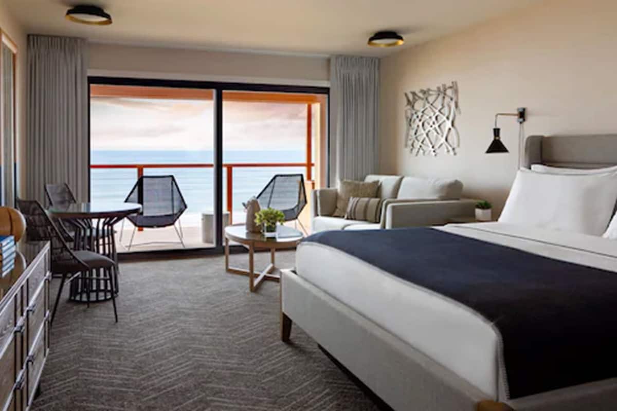 A hotel room with a large bed, a sofa, a desk with a chair, and a view of the sea through the balcony doors.