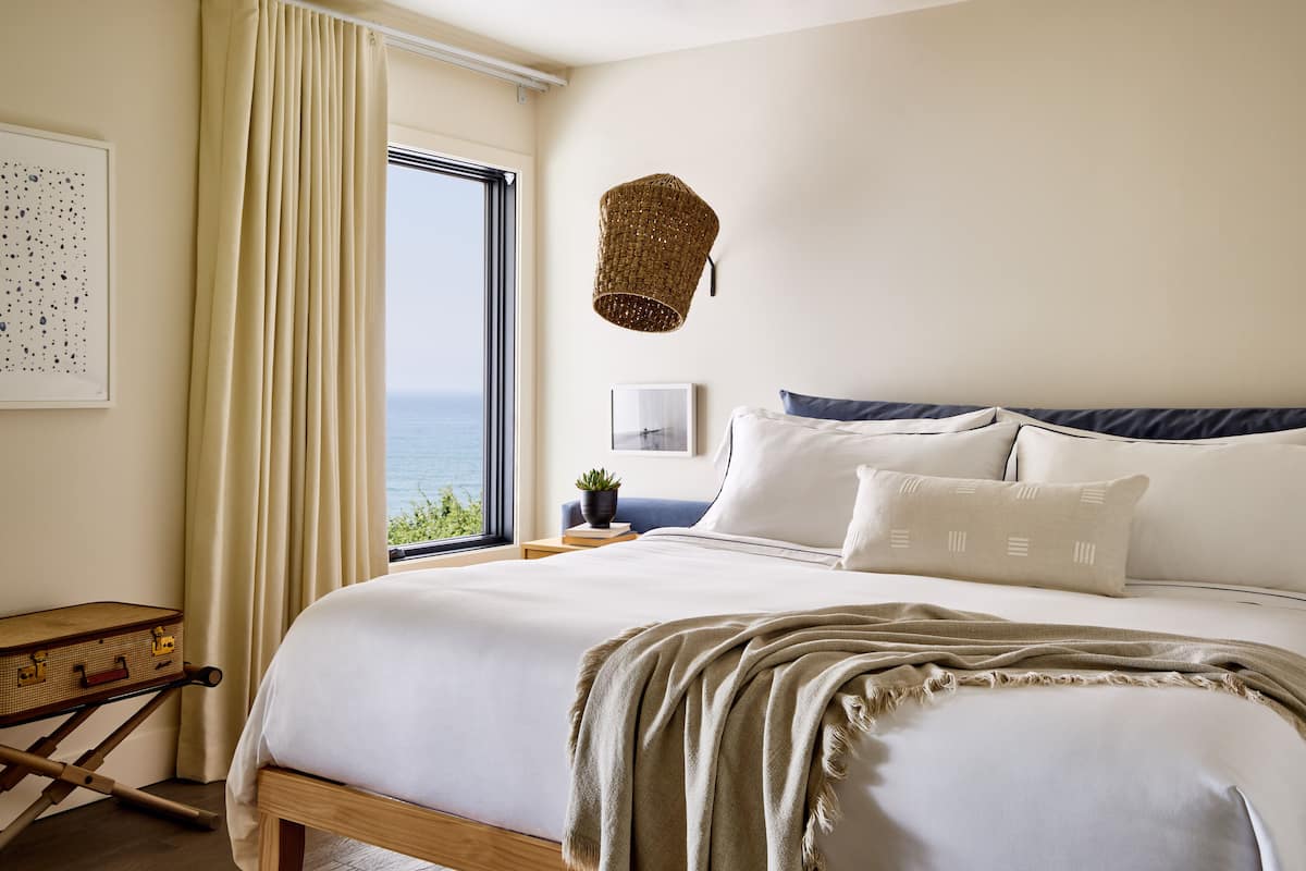 A bedroom with a large bed, a view of the sea, a wicker lamp, and a small table with a plant.
