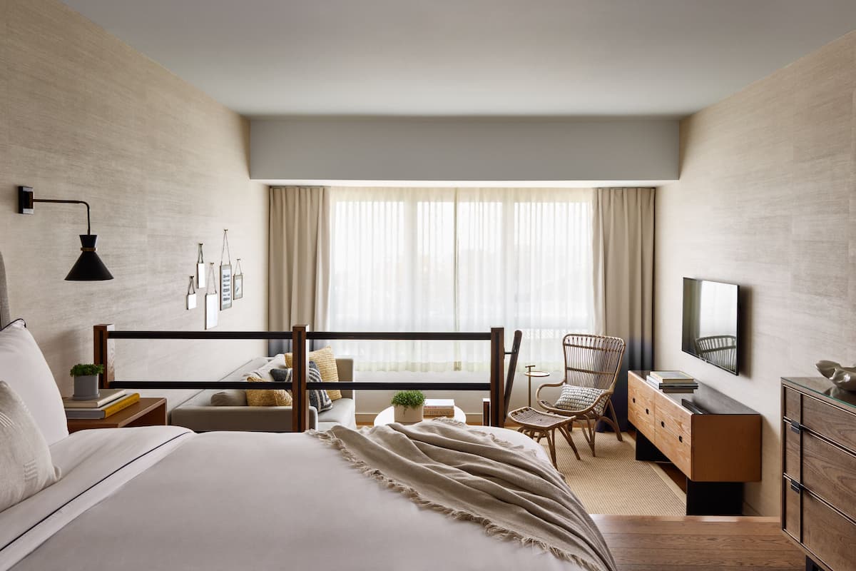 A modern hotel room with a bed, bedside table, lamp, chair, TV, dresser, and a large window with curtains.