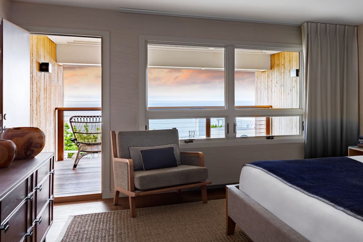 A hotel room with a large bed, a chair, a desk, and a view of the ocean through a window.