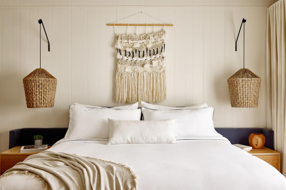 A neatly made bed with white linens and a blue headboard, flanked by two nightstands with woven baskets and a small plant.