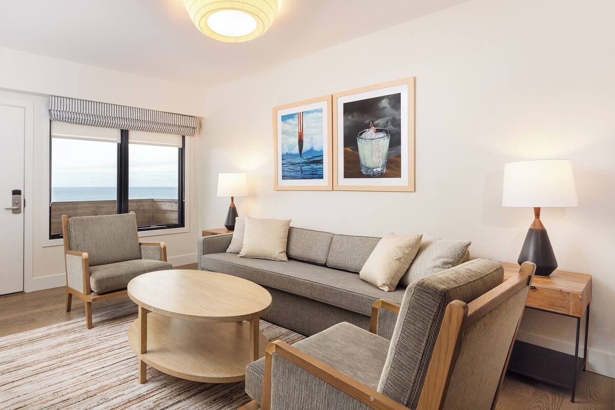 A modern living room with a sofa, two armchairs, a round coffee table, and a view of the ocean through large windows.