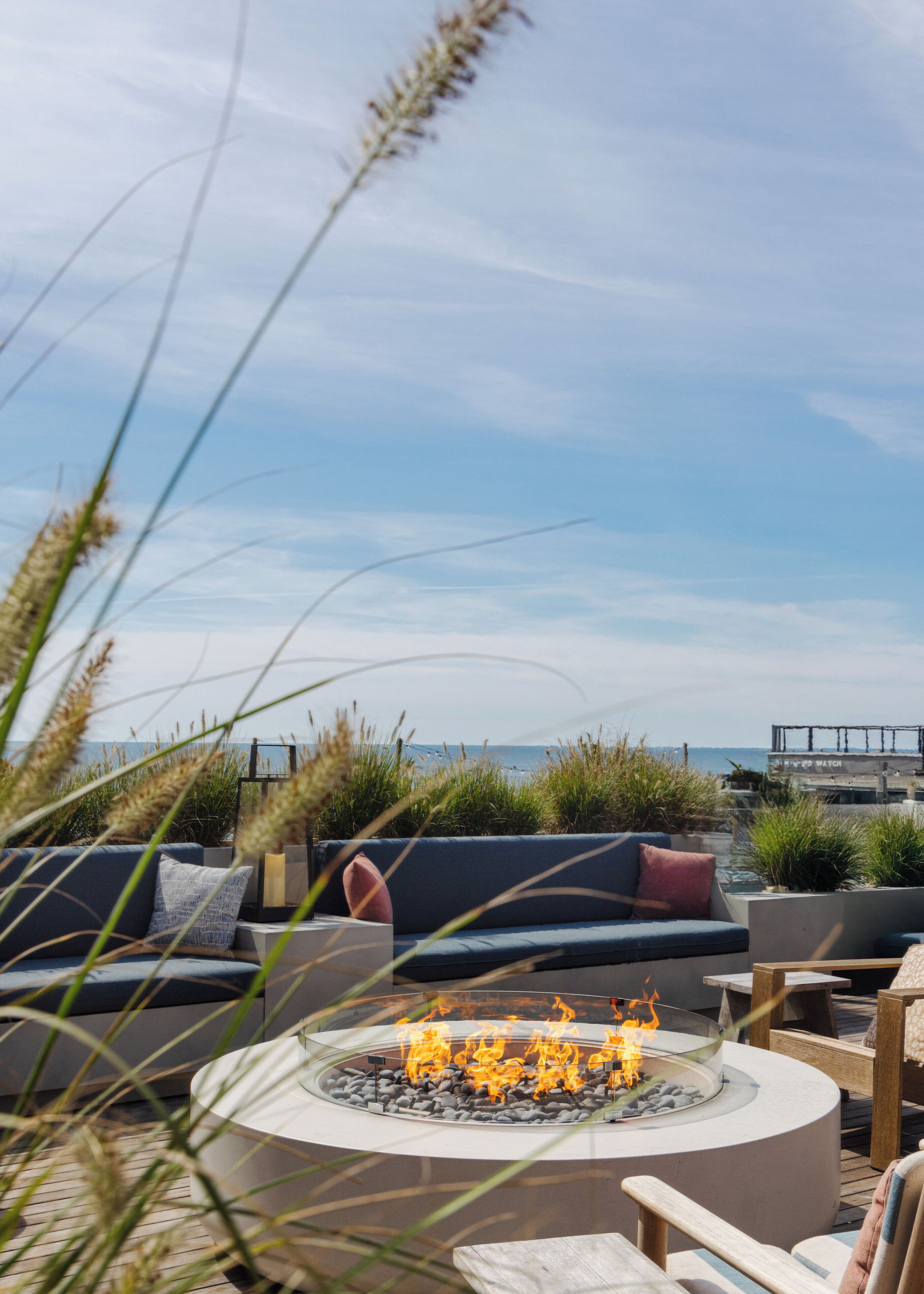 A circular outdoor seating area with a fire feature, surrounded by plants and overlooking a body of water.