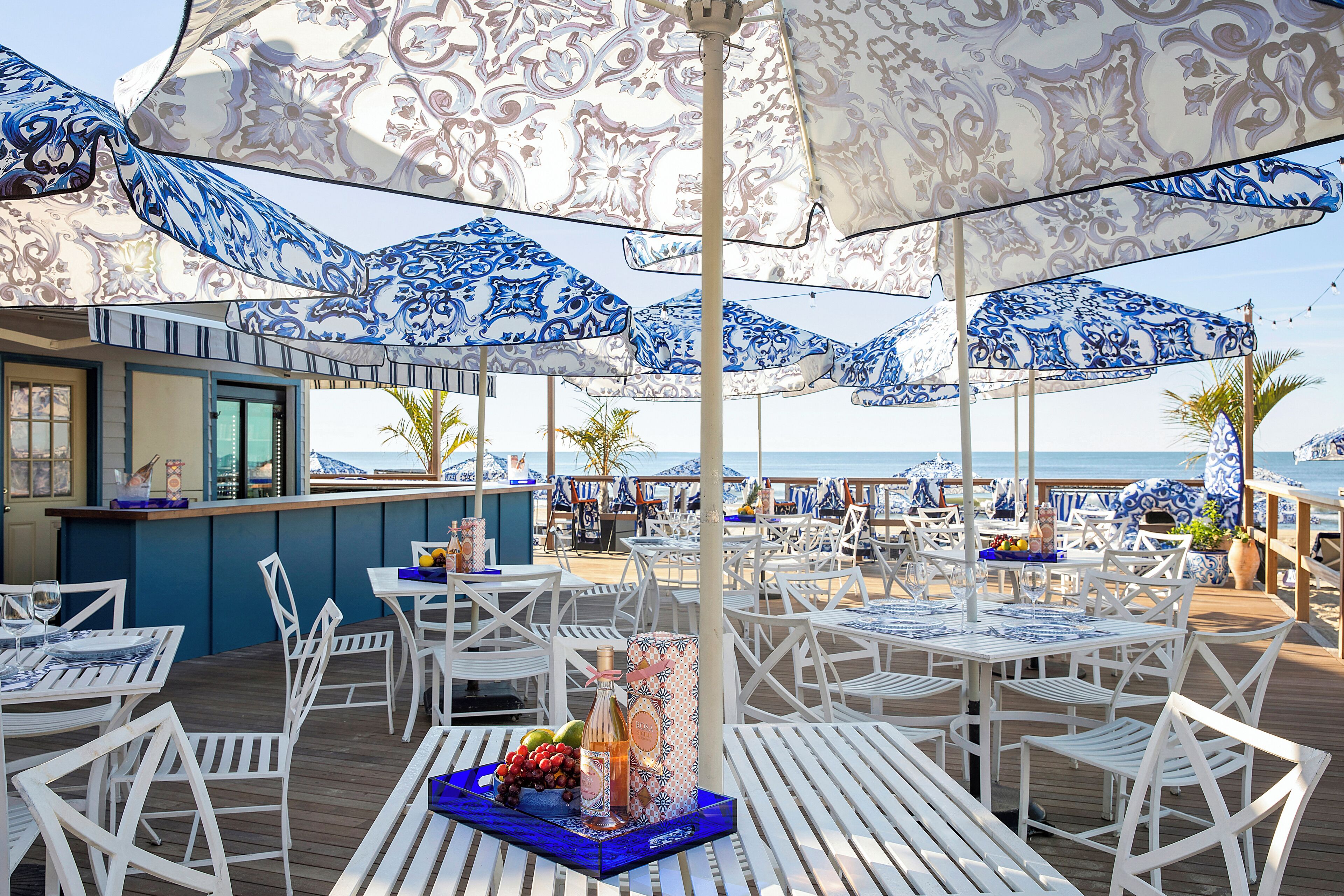 A beachside restaurant with white tables and blue patterned umbrellas, a bar area, and a view of the sea.