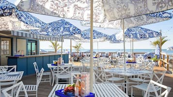 A beachside restaurant with white tables and blue patterned umbrellas, a bar area, and a view of the sea.