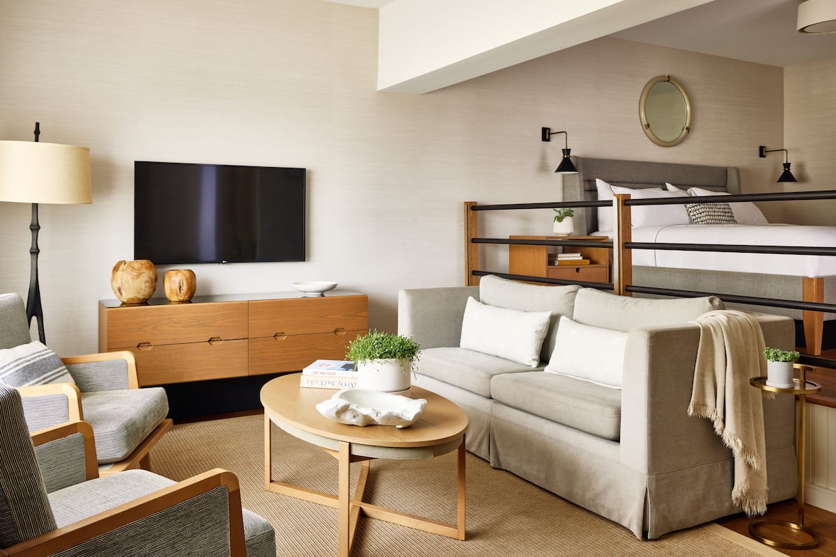 A modern hotel room with a sofa, a small round table, a TV, a bedside table with a lamp, and a bunk bed.