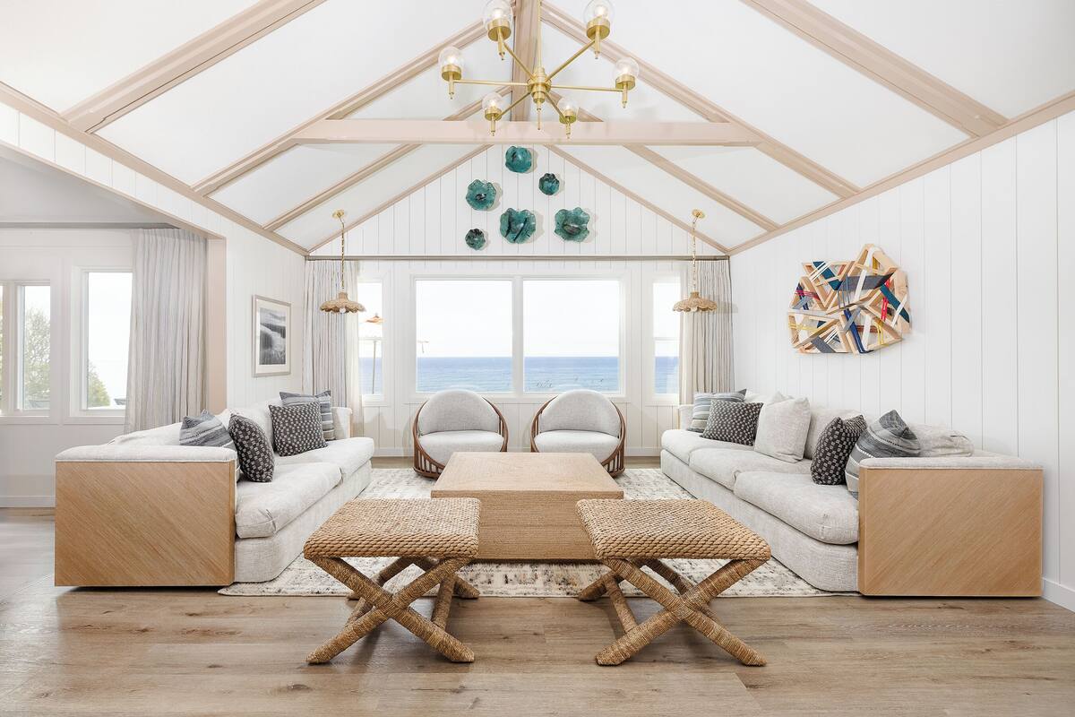 A spacious living room with a high ceiling, wooden beams, a large window with a view, two sofas, a coffee table, and a chandelier.