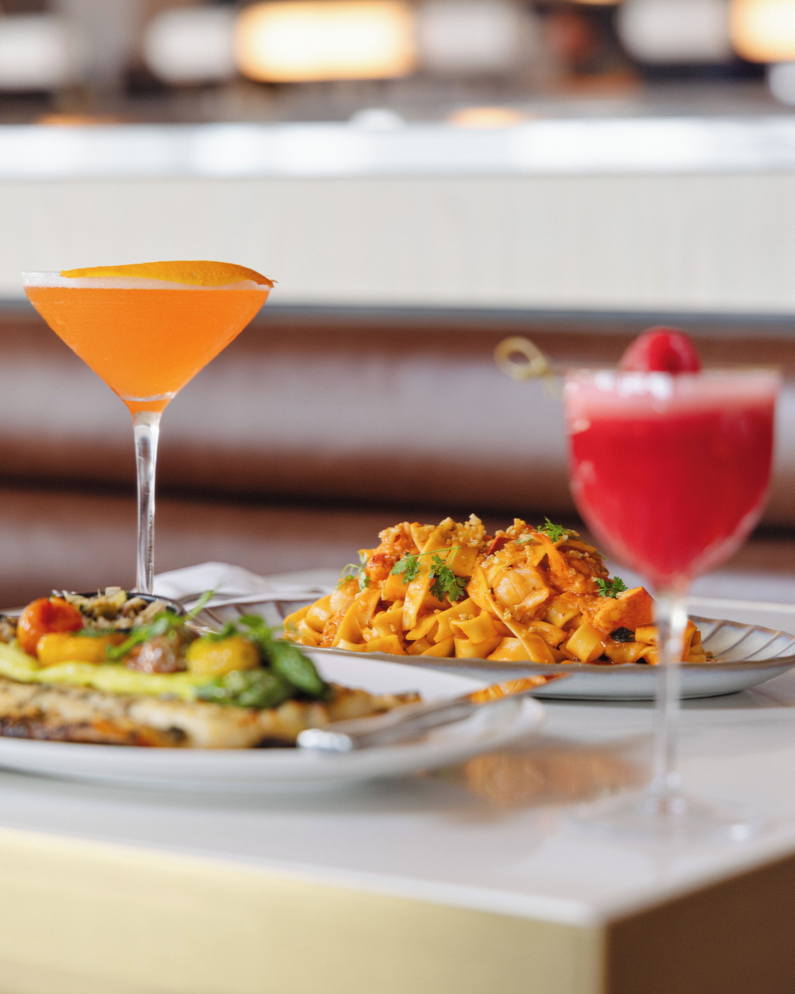 A dining table with two plates of pasta and two drinks, one orange and one red, in martini glasses.