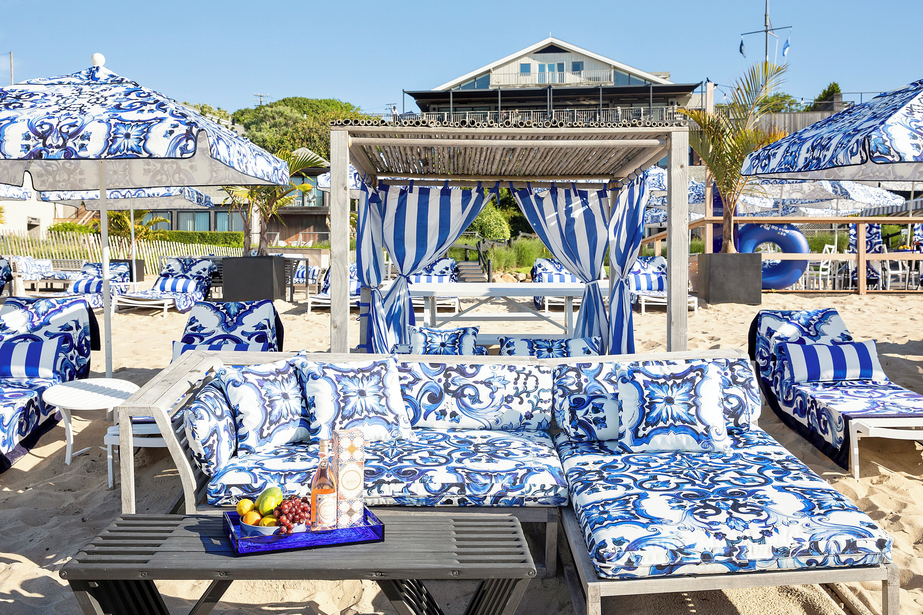 A beachside lounge area with blue and white patterned furniture, including sofas and chairs, and a table with a fruit bowl.
