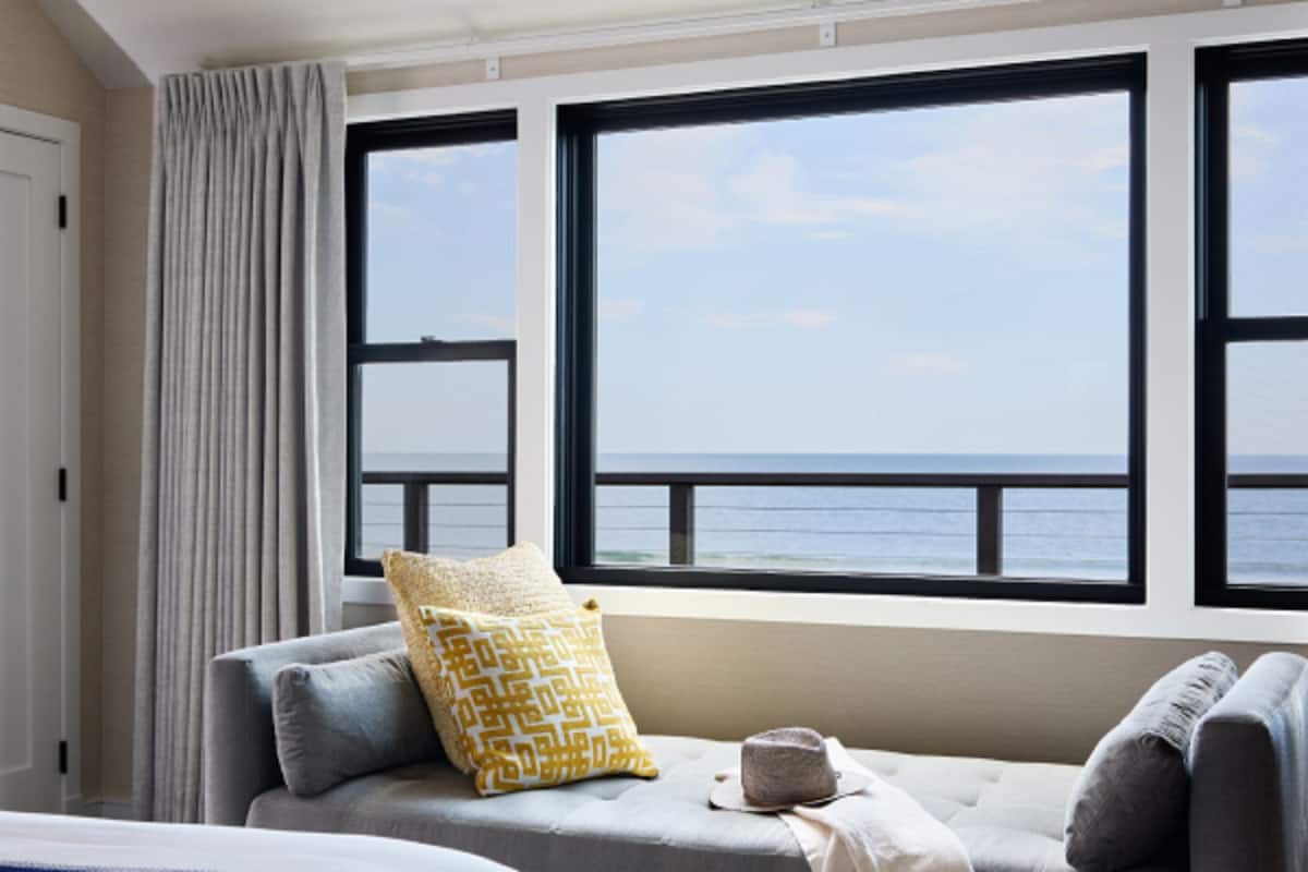 A room with a view of the sea through large windows, a sofa with cushions, and a curtain on the left.