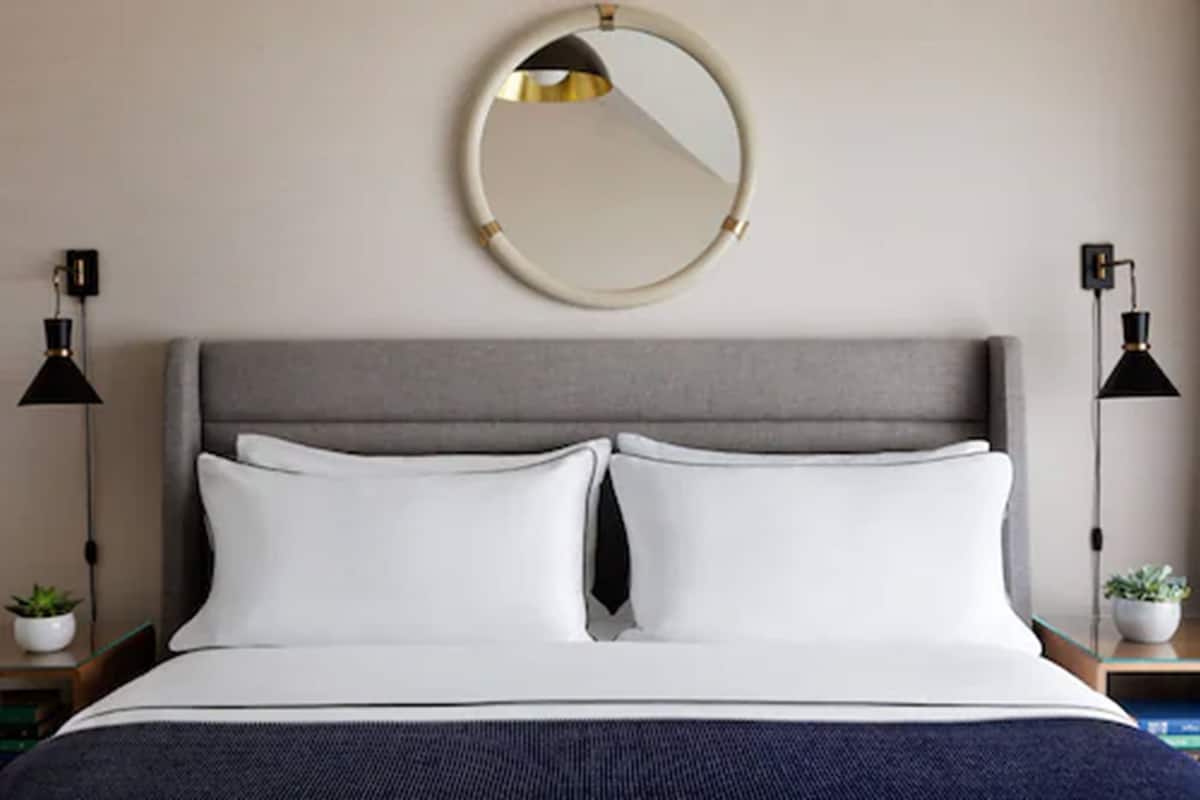A neatly made bed with white pillows and a dark blue throw, a round mirror on the wall, and two black floor lamps on either side.