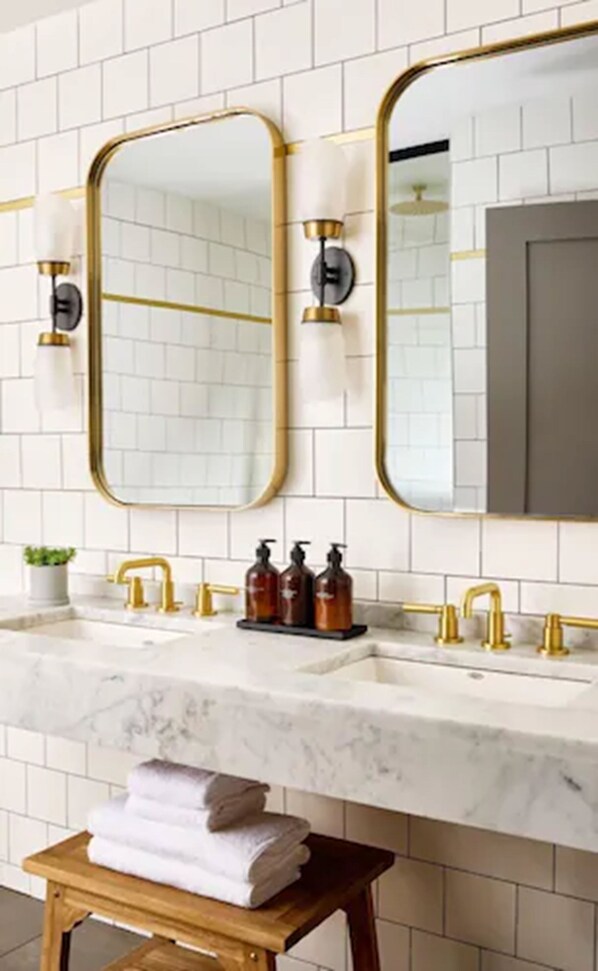 Un baño con paredes de azulejos blancos, encimera de mármol, detalles dorados y un espejo grande.