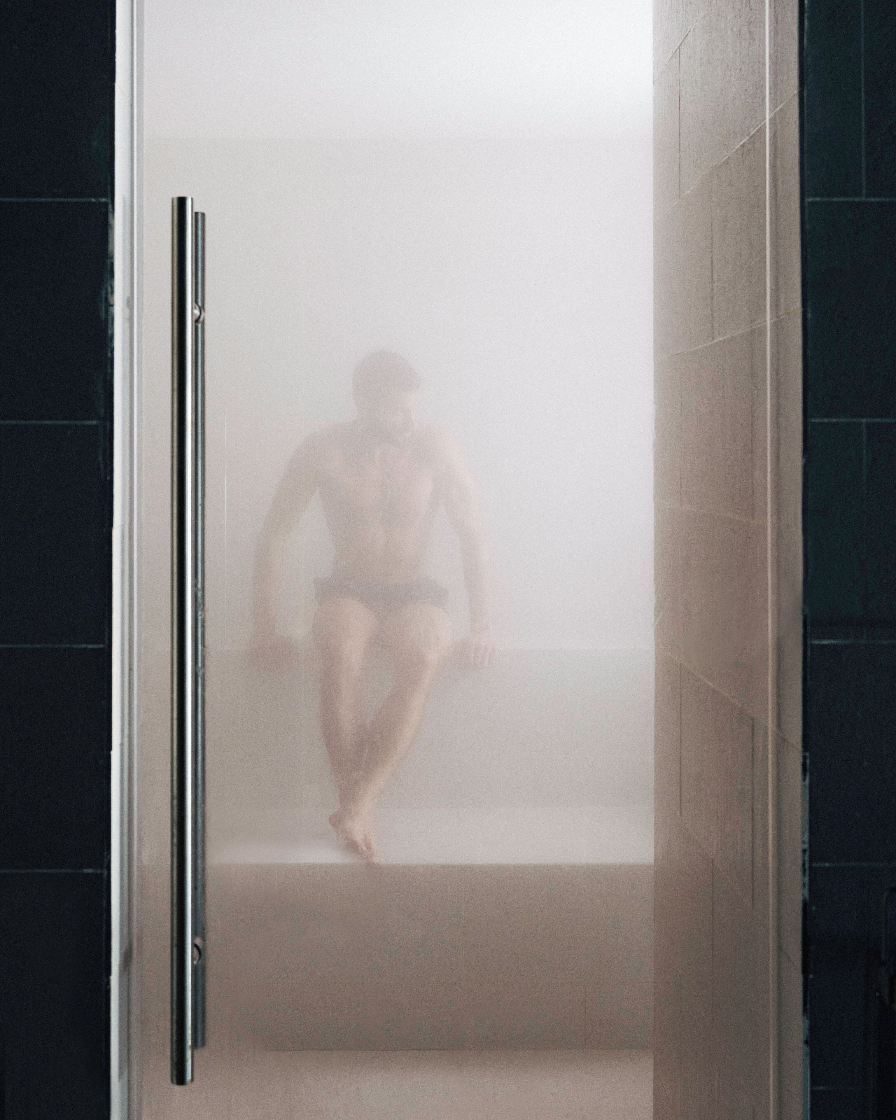 A person sitting on a staircase inside a bathroom with tiled walls and a glass door.