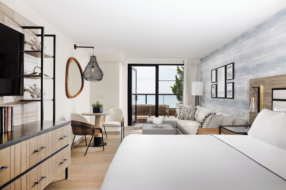 A modern hotel room with a large bed, a desk, a chair, a sofa, and a view of the sea.