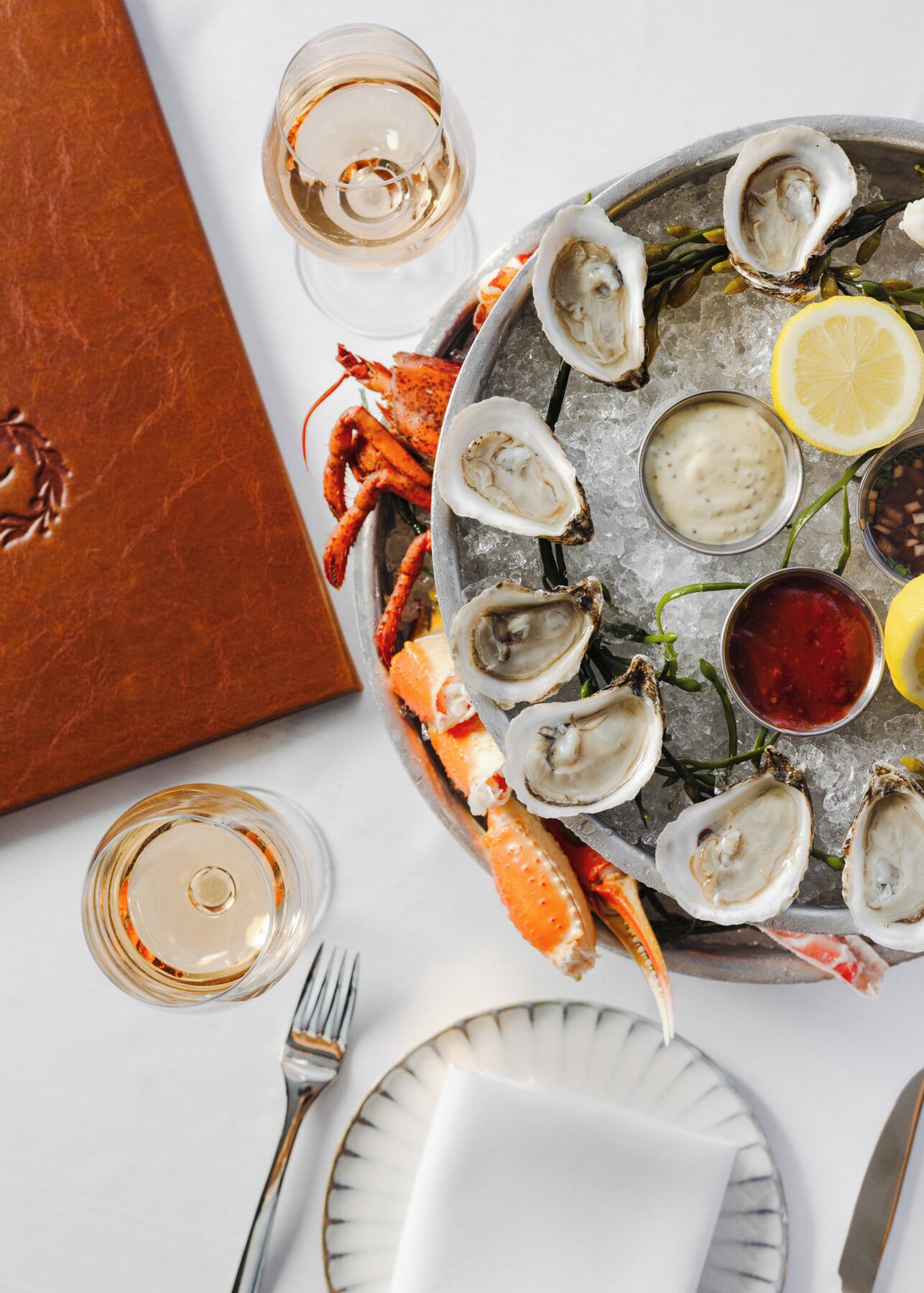 A seafood platter with oysters, lemon, and sauces on ice, accompanied by a glass of white wine and a lobster tail.