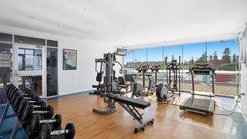 Un gimnasio con equipo de ejercicio, amplias ventanas y vista a edificios.
