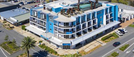 A modern hotel with a blue and white facade, multiple balconies, and a parking lot.
