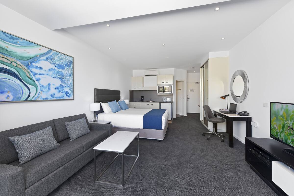 Ein modernes Hotelzimmer mit Bett, Sofa, Schreibtisch, Fernseher und einer Küchenzeile.