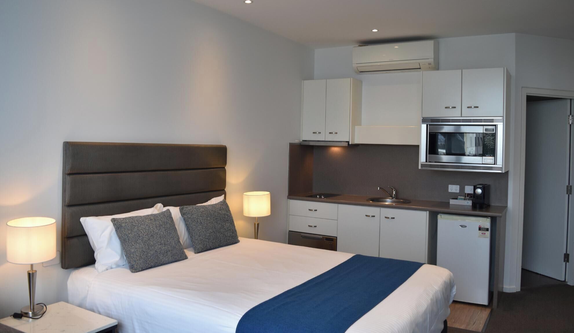 Habitación de hotel tipo estudio con cama, lámparas de noche, kitchenette con microondas y un refrigerador pequeño.