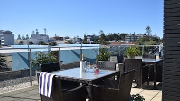 Une terrasse extérieure aménagée avec des meubles en osier, une table en verre et une vue sur le paysage urbain.
