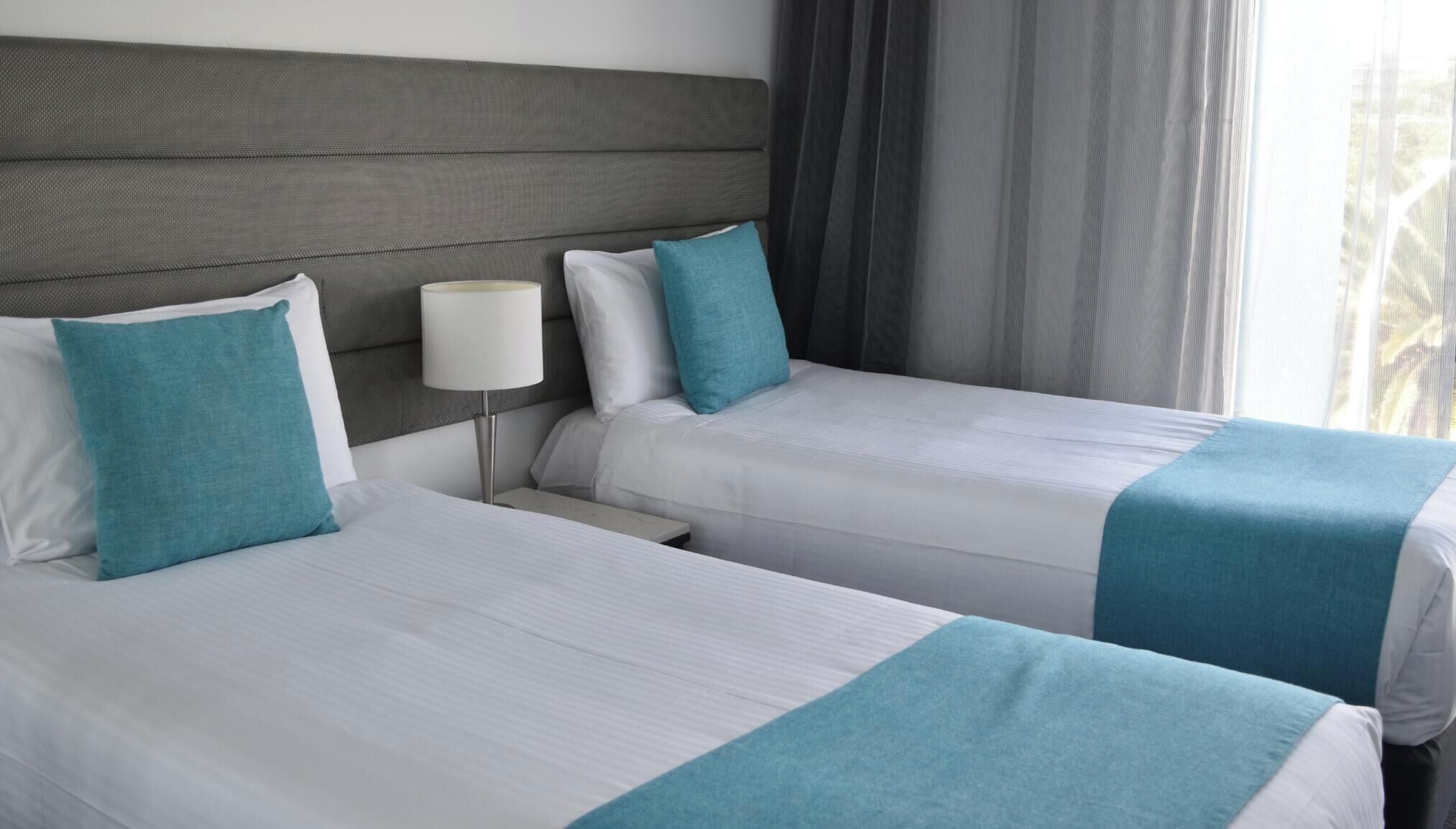 A 2nd bedroom with two beds, each with white bedding and teal accents, a bedside table with a lamp, and a view of the outdoors through a window with curtains.