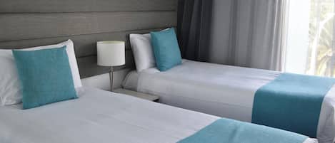Een tweede slaapkamer met twee bedden, elk met witte beddengoed en teal accenten, een nachtkastje met een lamp, en uitzicht op het buitenlands landschap door een raam met gordijnen.