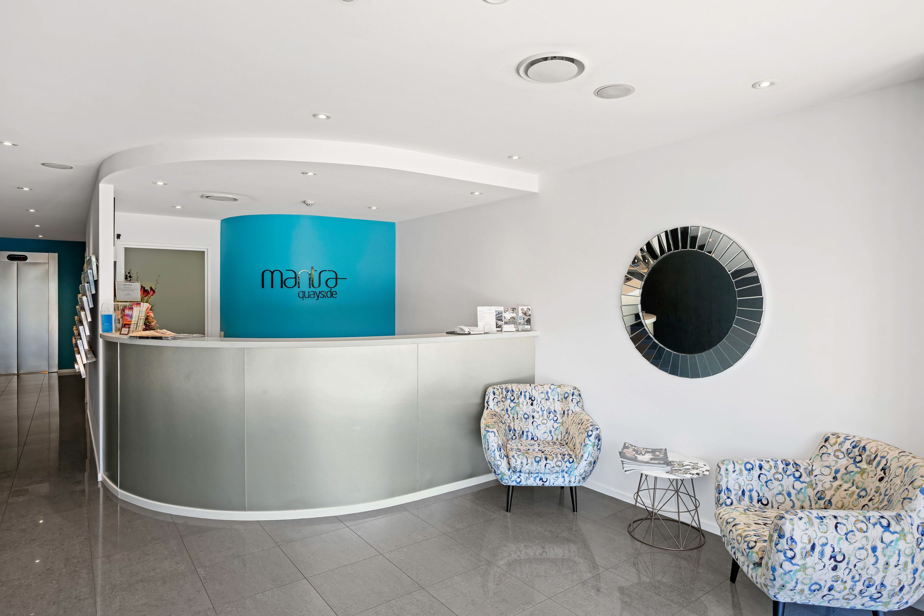 A reception area with a curved counter, a blue accent wall, two patterned chairs, and a round mirror on the wall.