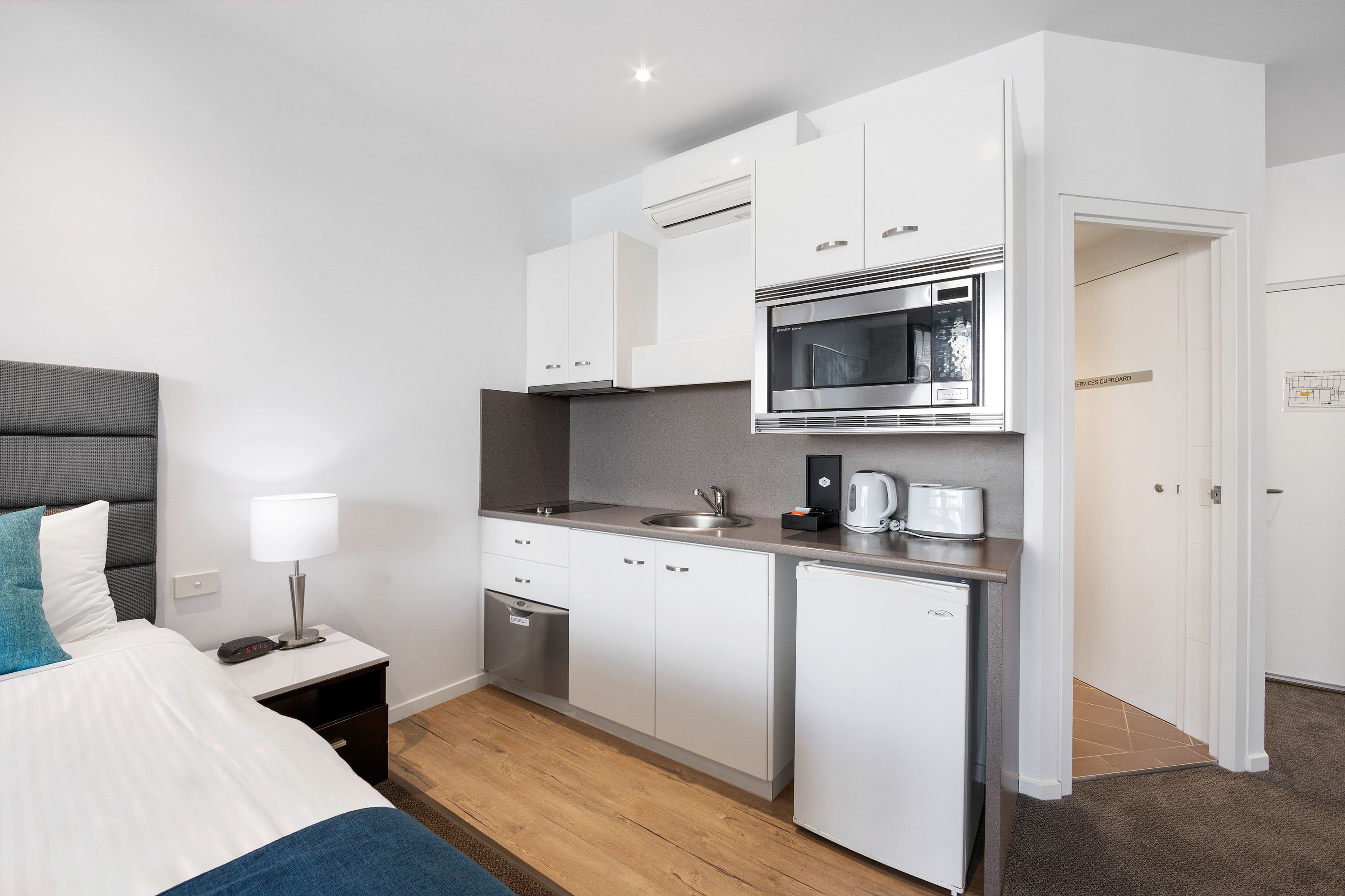 A compact hotel room with a kitchenette, a bed with blue bedding, a bedside table with a lamp, and a small refrigerator.