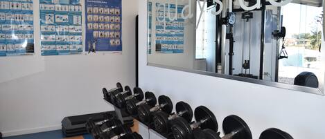A gym with dumbbells on racks, exercise posters on the wall, and a large mirror reflecting gym equipment.