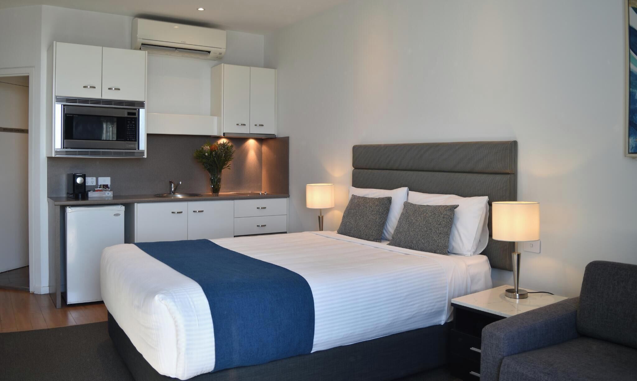 A modern hotel room with a bed, bedside tables, a desk, and a kitchenette.
