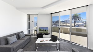 Una sala moderna con un sofá gris, una mesa de centro con una computadora portátil y una revista, un televisor sobre un mueble negro y grandes puertas correderas de vidrio que dan a un balcón con vistas a edificios y palmeras.