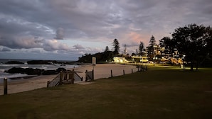Vista costera con playa, valla de madera y un edificio con ventanas iluminadas.