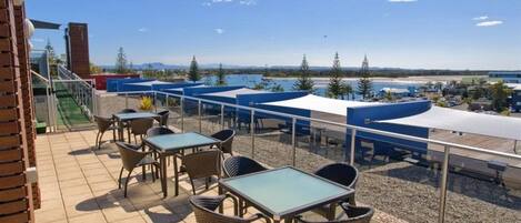 A terrace with outdoor seating, a view of the sea, and a building in the background.
