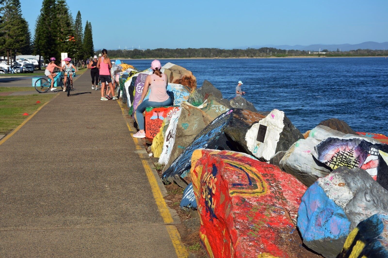 A pathway along a waterfront with colorful rocks and a few cyclists.