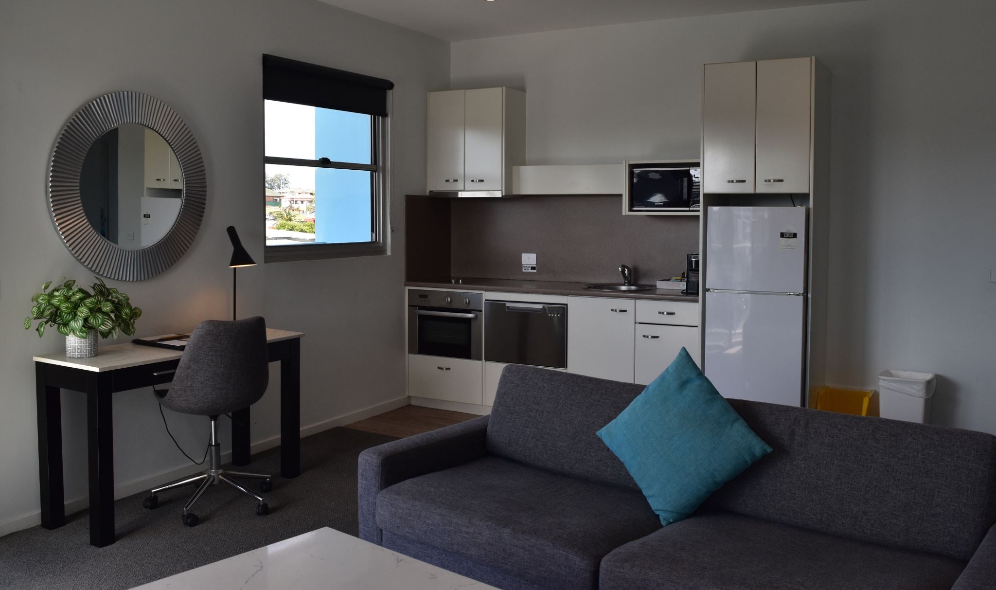 A 2 bedroom living room/kitchen with a modern design, including a sofa, a desk, a chair, a mirror, cabinets, and a kitchen with appliances.
