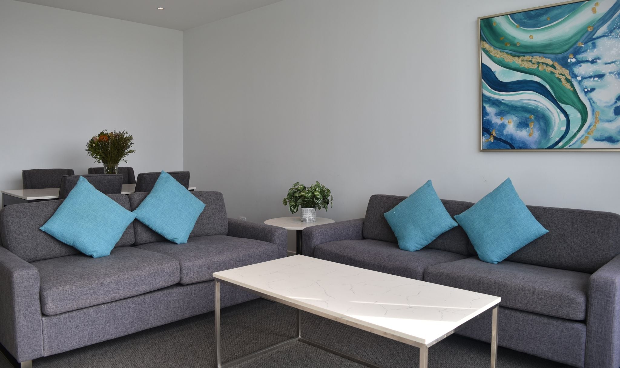 A 1-bedroom living room with two grey sofas, a white coffee table, a small round table with a plant, and a large abstract painting on the wall.