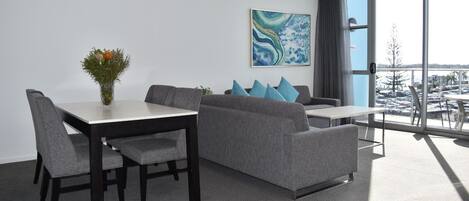 A 1-bedroom living room with a dining table, grey sofa, and a large window offering a view of the parking lot.