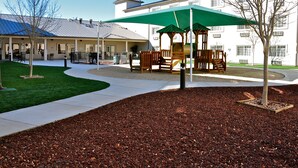 A playground with a green canopy, surrounded by a landscaped area with mulch and trees.