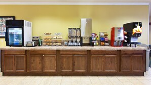 A kitchen area with a marble countertop, wooden cabinets, a refrigerator, a microwave, a coffee machine, and various food items.