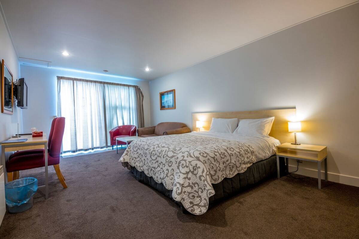 Habitación de hotel con una cama, un escritorio, una silla, una silla roja, una lámpara y un ventanal con cortinas.