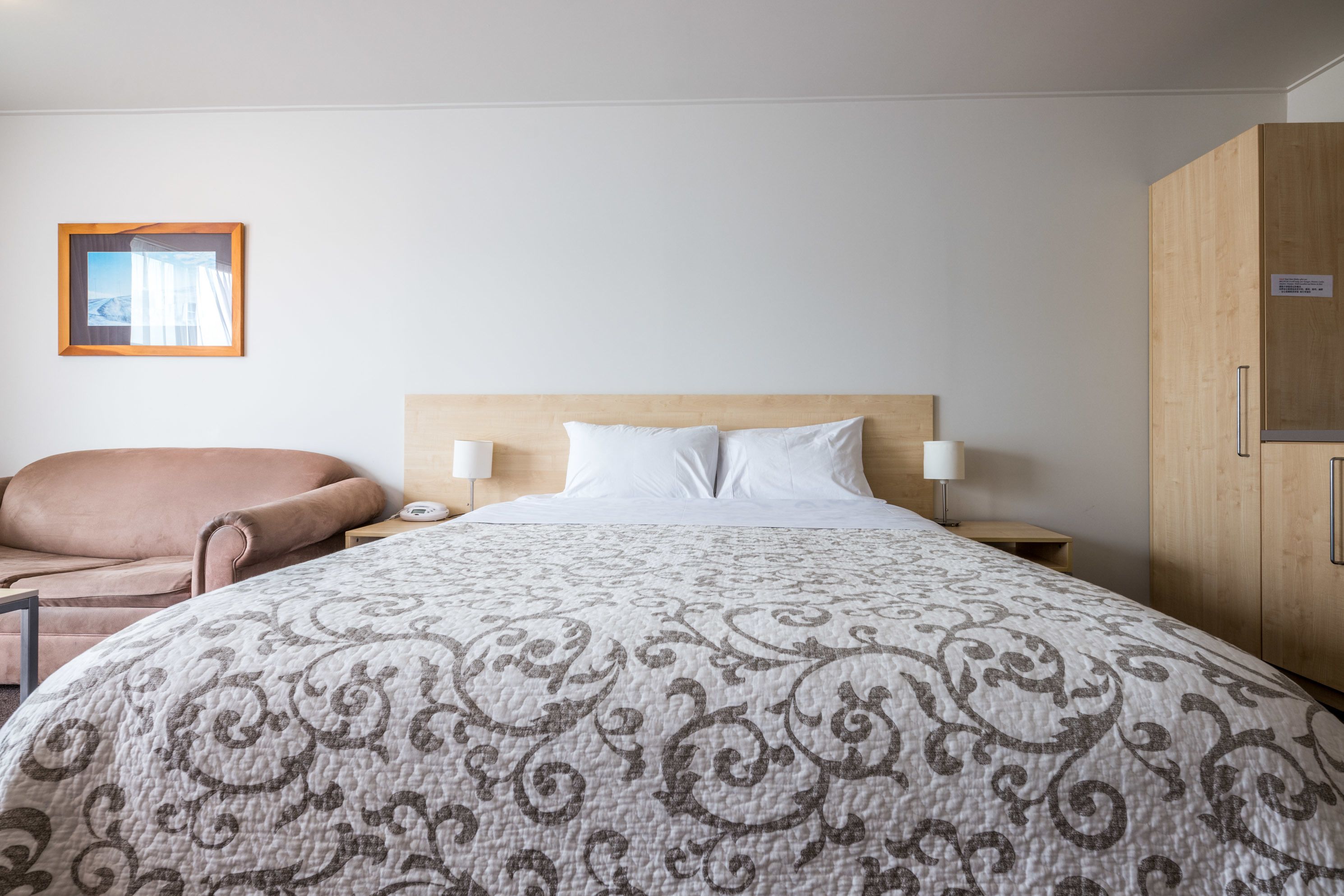 A hotel room with a large bed, a wooden headboard, a nightstand with a lamp, a chair, and a wardrobe.