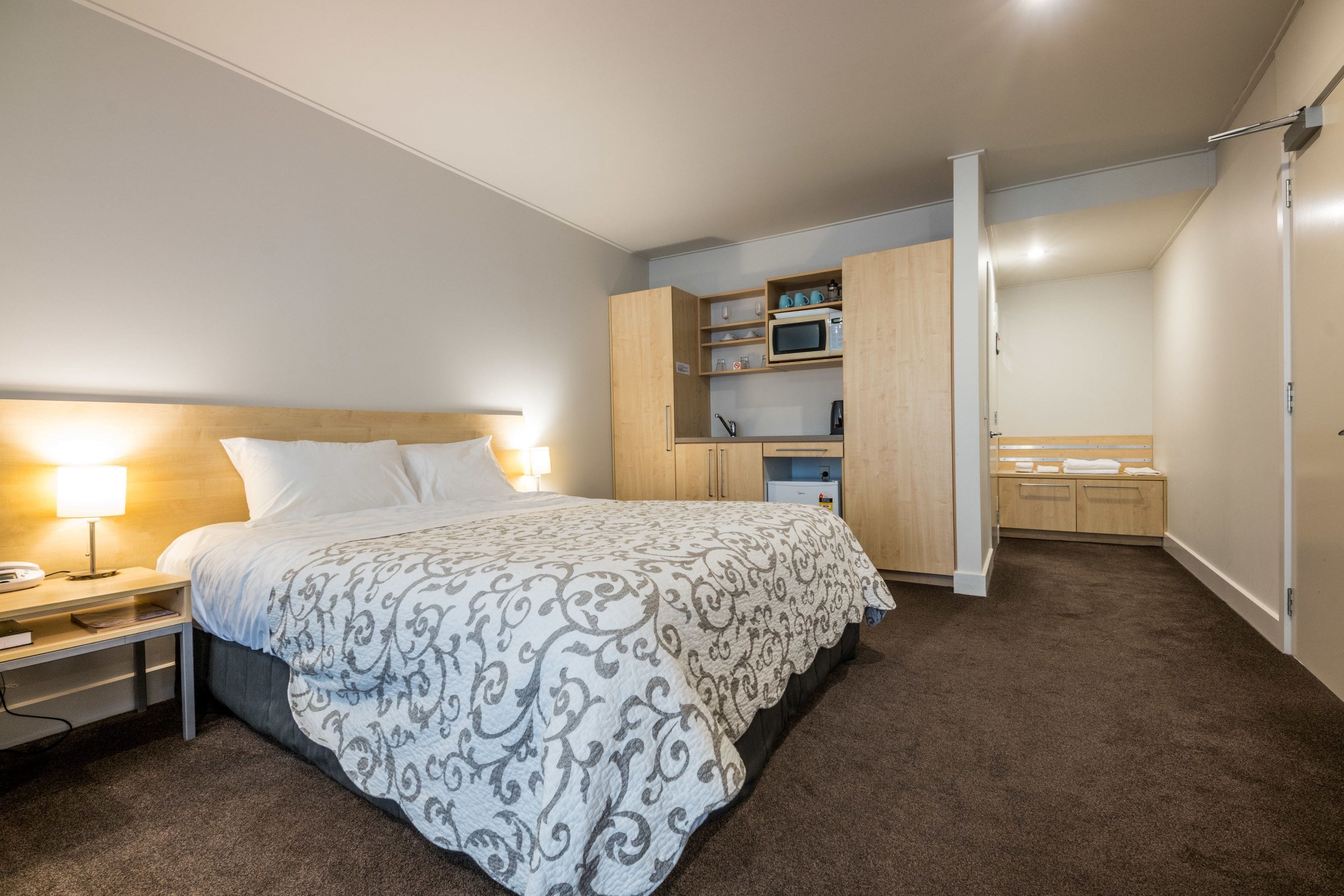 A bedroom with a bed, bedside table, lamp, and a built-in kitchenette with a microwave and storage cabinets.