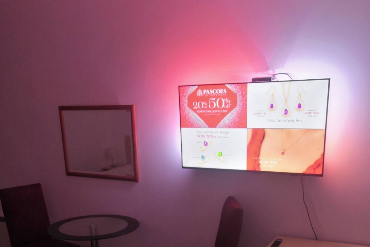 Habitación de hotel con cama, una mesa redonda, dos sillas, un televisor mostrando un anuncio de joyería y un cuadro en la pared.