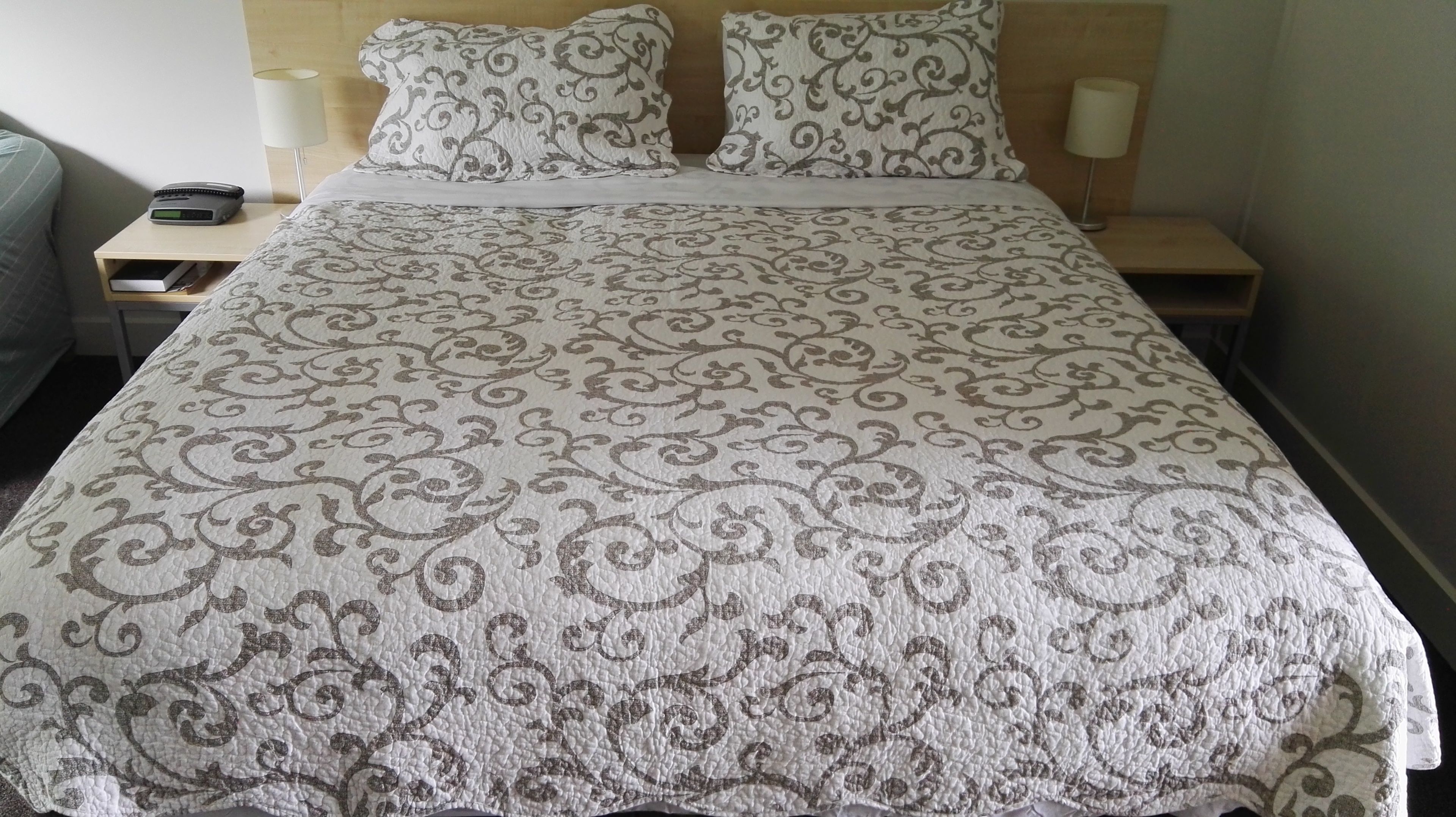 A neatly made bed with a patterned bedspread, two bedside lamps, and a bedside table with a phone and books.
