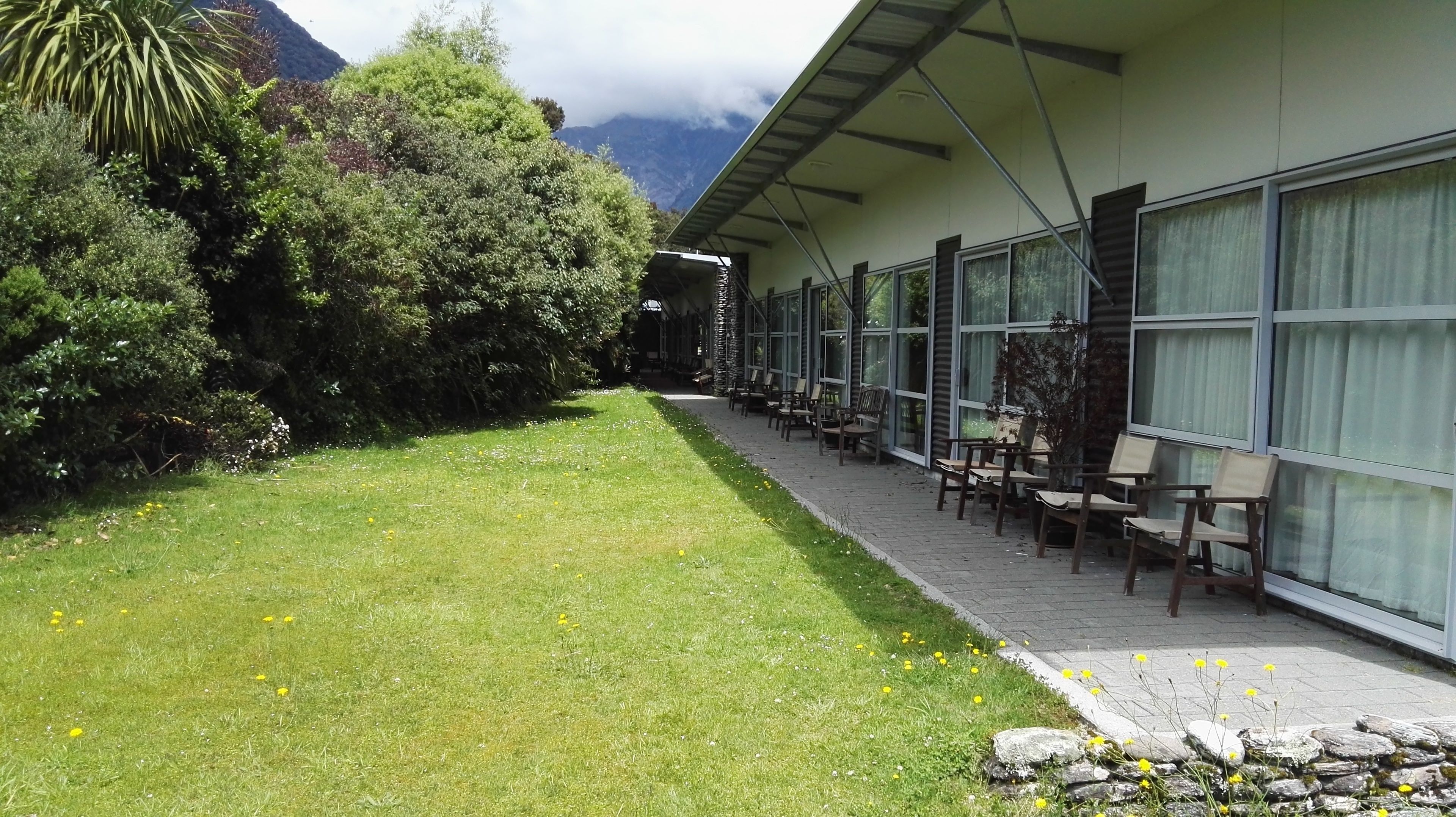 A hotel with a covered outdoor seating area, a grassy lawn, and a mountain backdrop.
